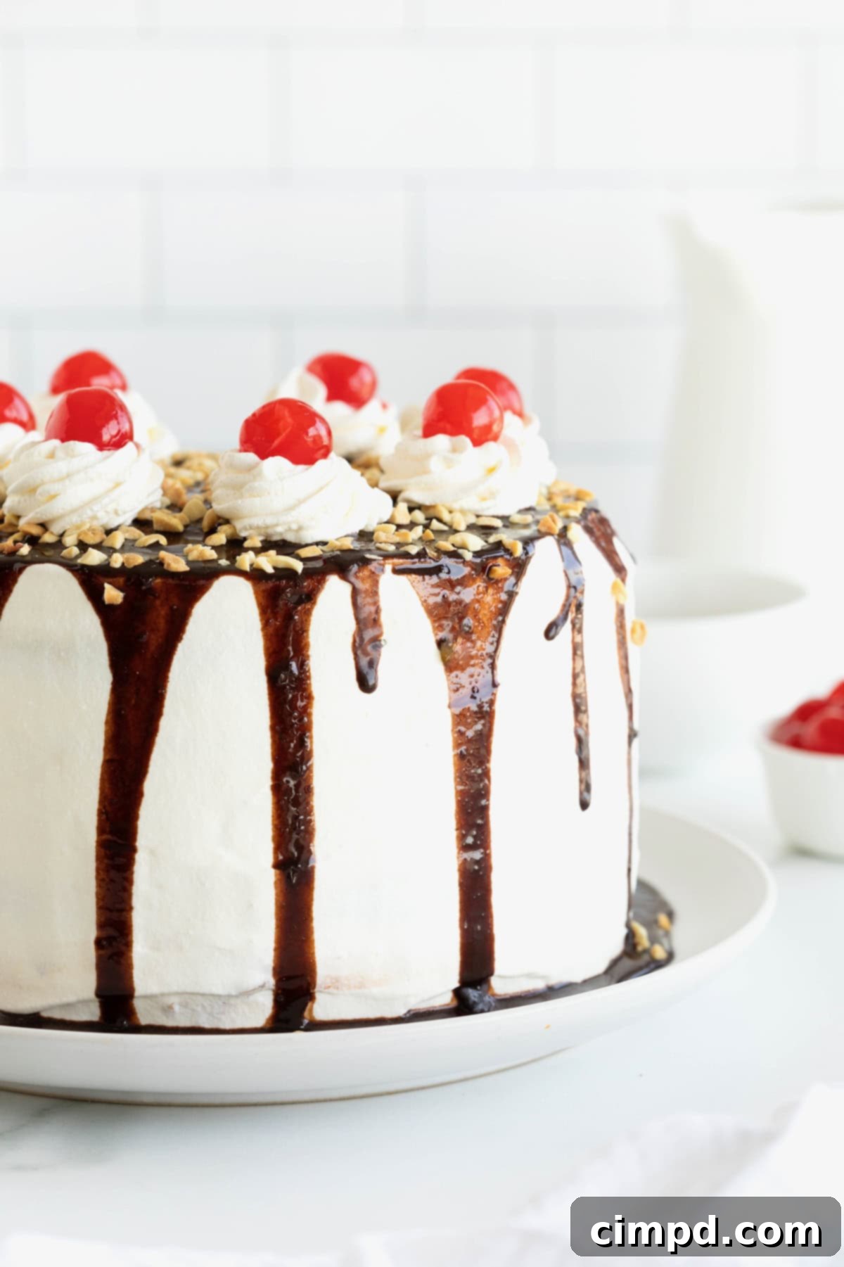 Banana Split Dream Cake 3 A stunning Banana Split Cake, artfully topped with bright red cherries, presented on a graceful white cake plate.