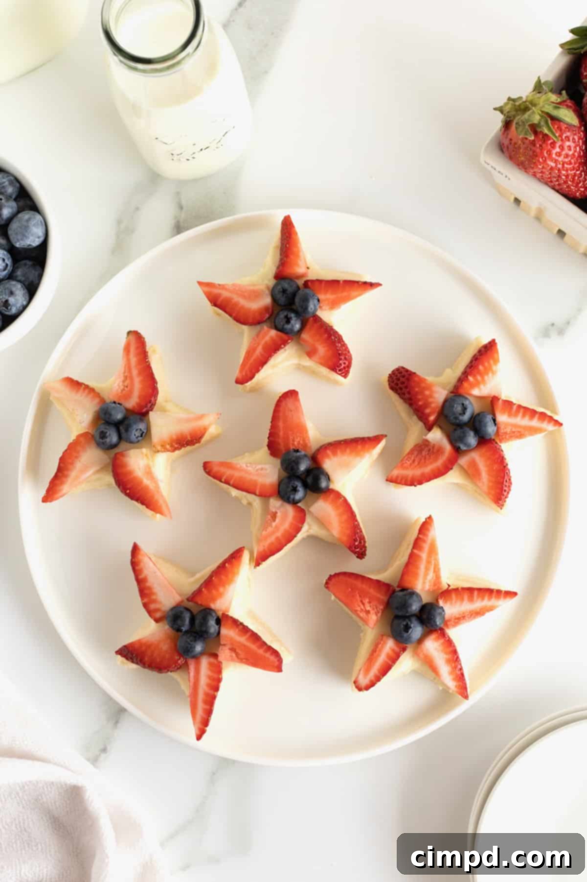 Star-Spangled Celebration Cookies 3 A beautiful round, white serving platter showcasing an array of star shaped shortbread cookies, each generously topped with strawberry slices and fresh blueberries.