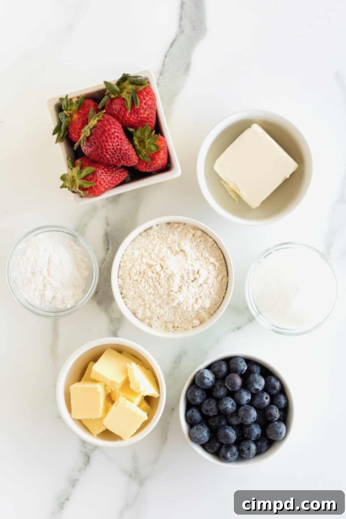 Star-Spangled Celebration Cookies 6 An organized display of ingredients for Fourth of July Star Cookies, including strawberries, blueberries, cream cheese, butter, sugar, and flour in small glass dishes on a pristine marble counter.