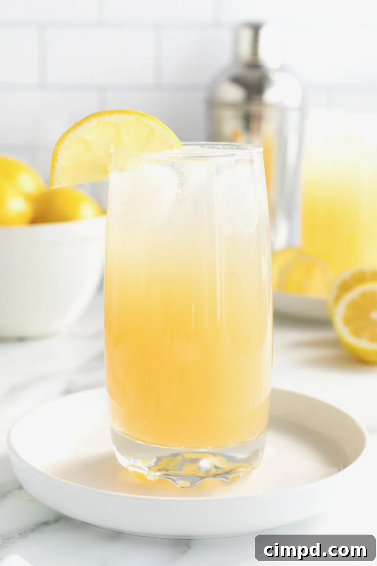 A tall glass of Zesty Lemonade Cocktail, garnished with a lemon slice, sits on a white rimmed tray on a white marble counter. In the background, a white bowl overflows with fresh lemons, suggesting the vibrant citrus notes of the drink.