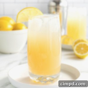A tall glass of Zesty Lemonade Cocktail on a rimmed white tray on a white marble counter. In the background is a white bowl filled with lemons.