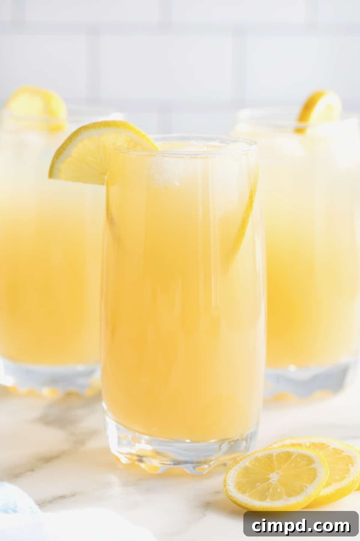 Three tall glasses of Zesty Lemonade Cocktail, each beautifully garnished with a vibrant lemon wedge, sitting on a pristine white surface, ready to be enjoyed.