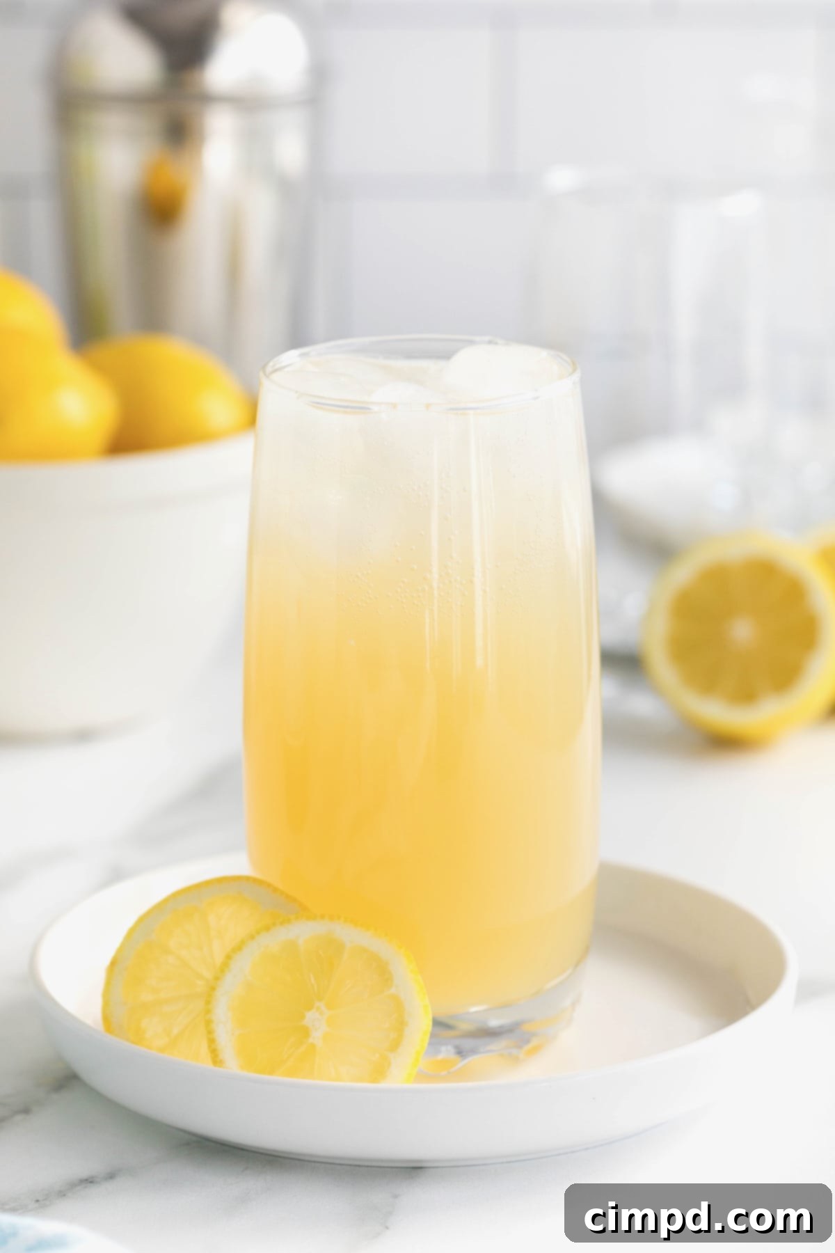 A single tall glass of the refreshing Zesty Lemonade Cocktail resting on a white rimmed tray on a white marble counter. The background features a bowl filled with bright yellow lemons, highlighting the key ingredient of the drink.
