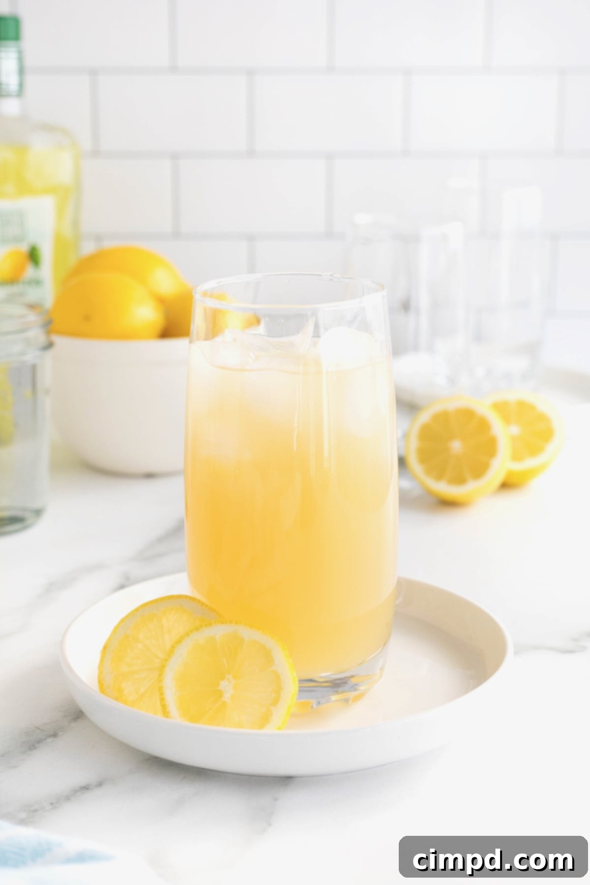 A tall glass of Zesty Lemonade Cocktail, complete with ice and a lemon garnish, rests elegantly on a white rimmed tray on a white marble counter. In the background, a white bowl filled with bright lemons reinforces the fresh citrus theme.