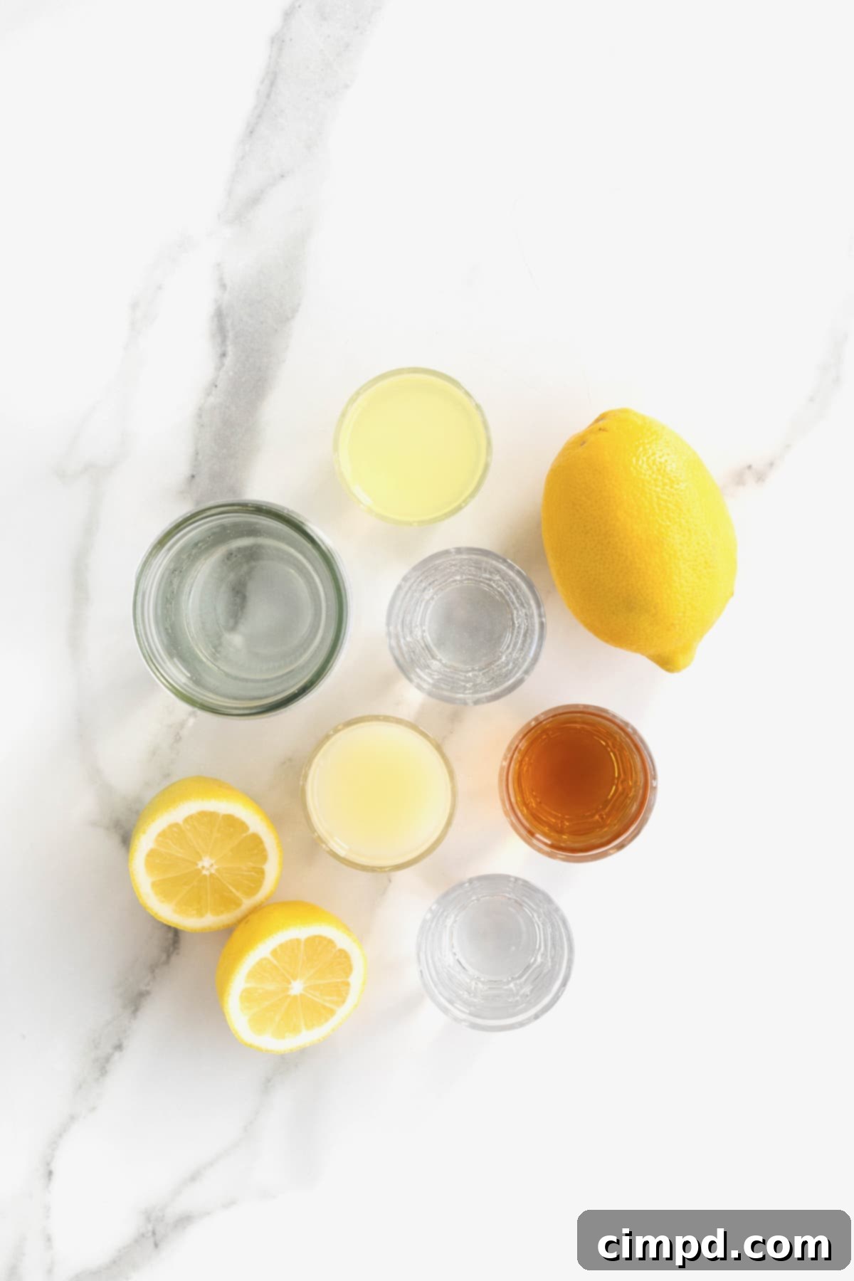 All the ingredients laid out on a white marble counter for making the Zesty Lemonade Cocktail: bottles of vodka, limoncello, and chile liqueur, fresh lemons, simple syrup, and sparkling water.