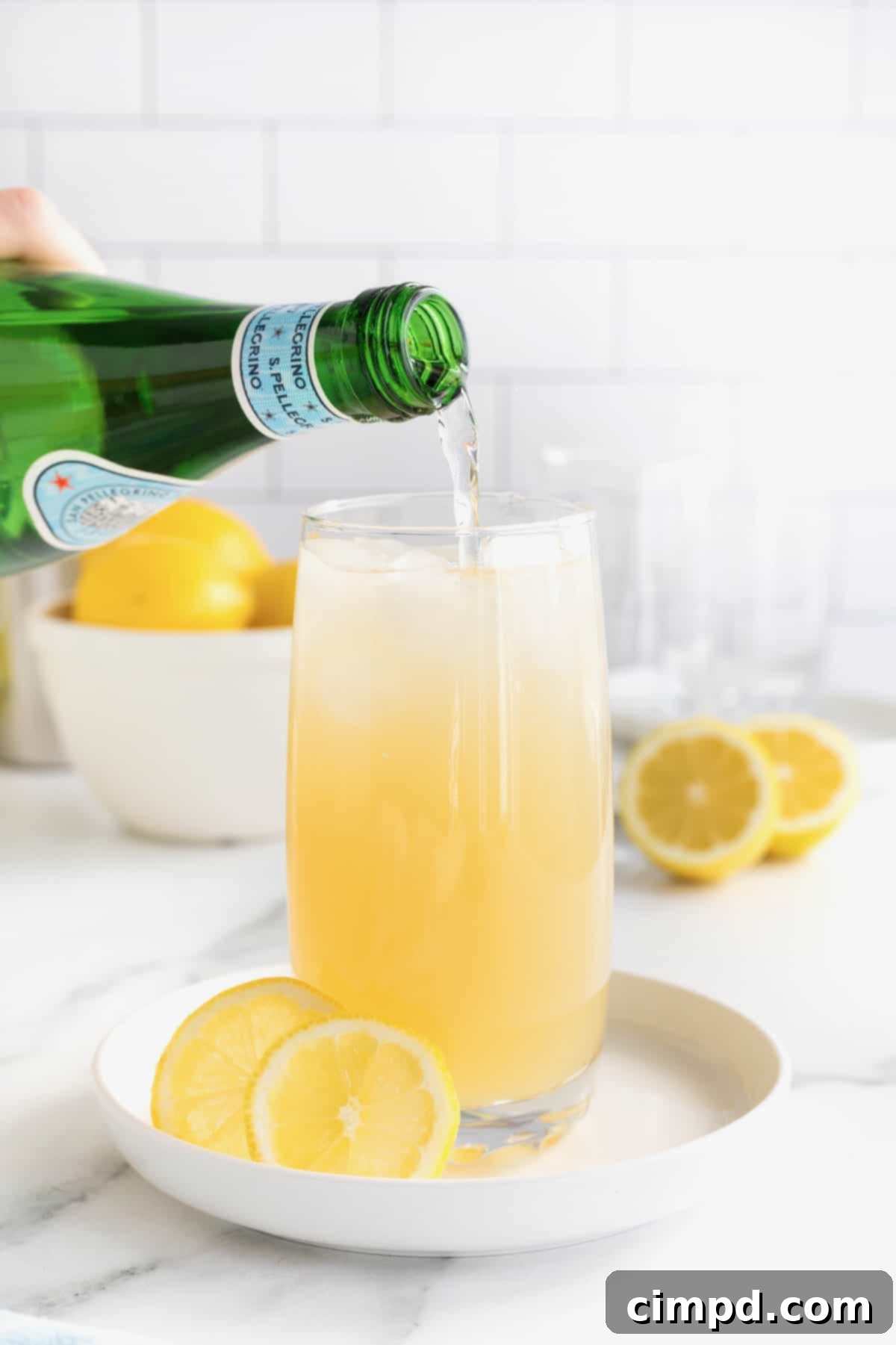 A hand pouring Pellegrino sparkling water into a glass of Zesty Lemonade Cocktail already filled with ice and a lemon garnish, completing the drink.
