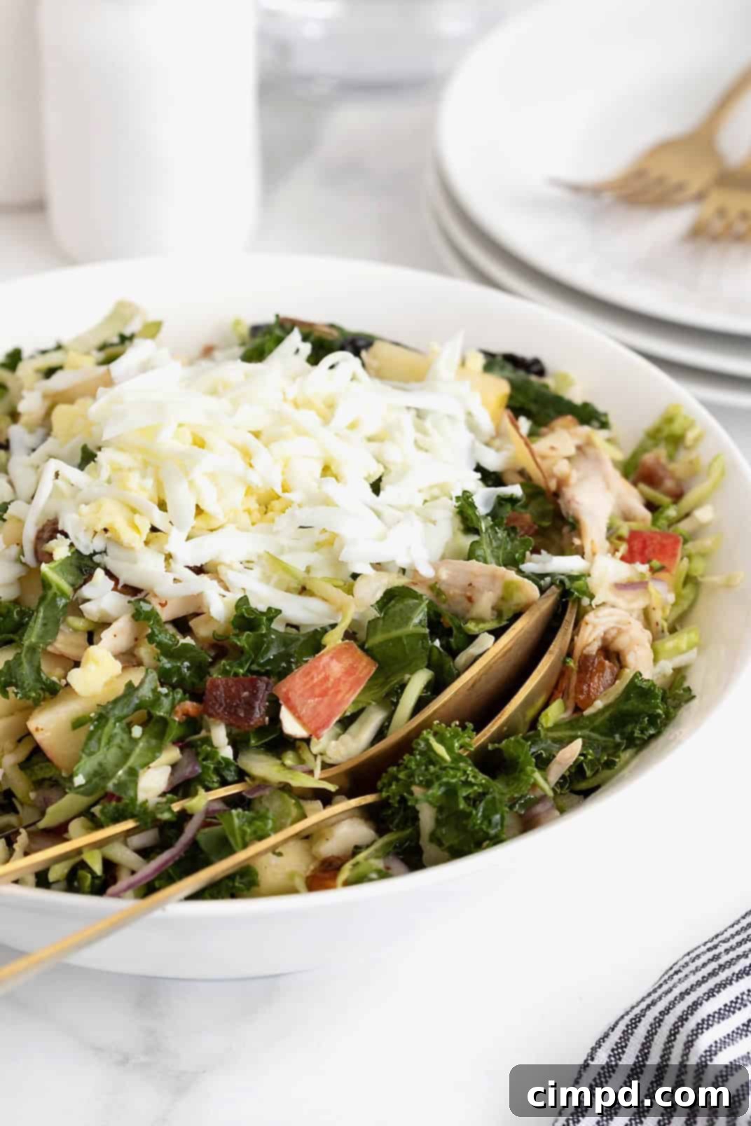 A large white serving bowl of kale salad with two gold toned spoons inside.