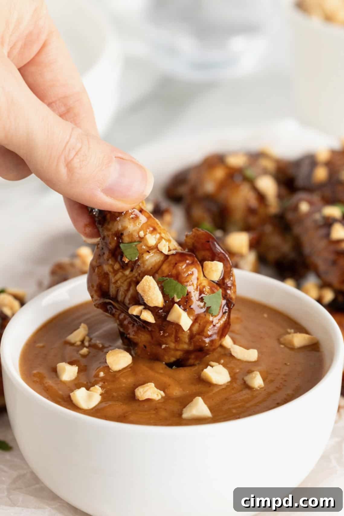 A close-up shot of a Baked Crispy Asian Chicken Wing being dipped into a creamy peanut sauce, showcasing its vibrant glaze and inviting texture.