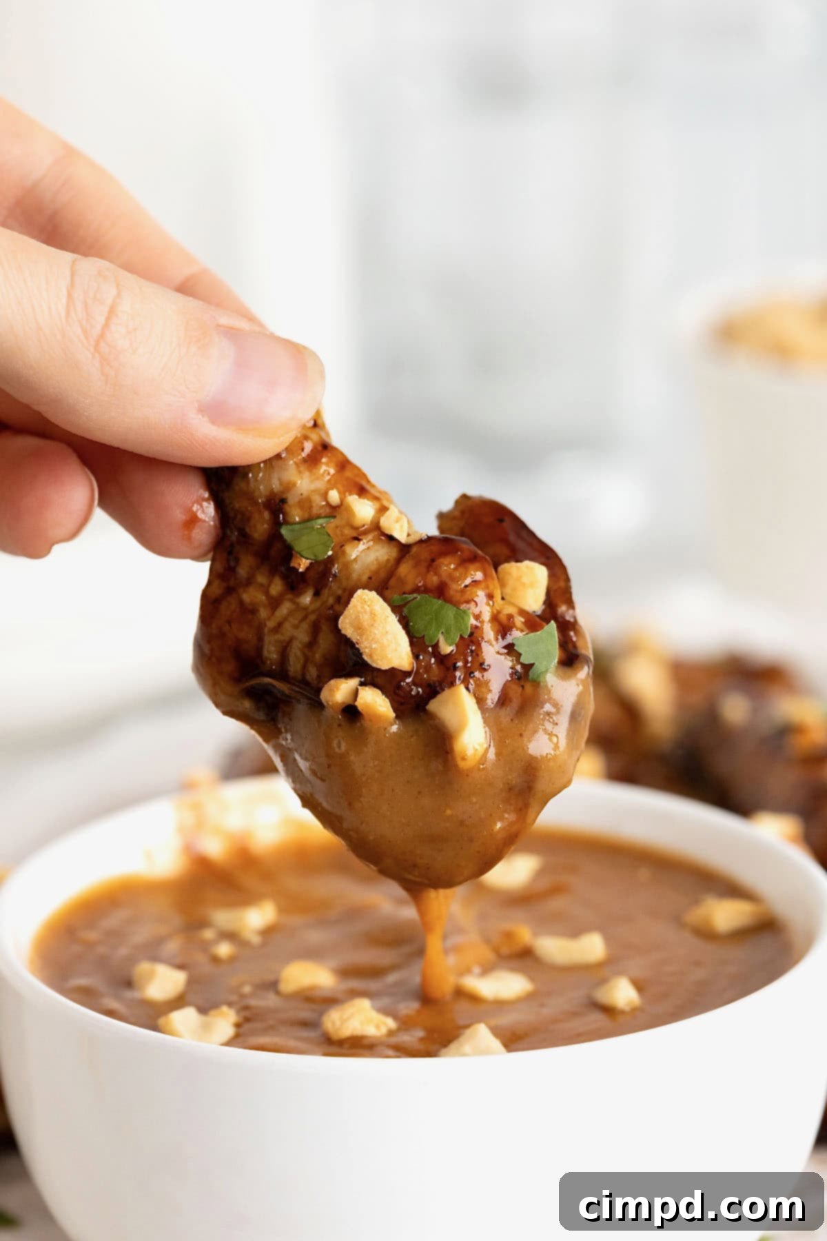A close-up of a perfectly glazed Baked Crispy Asian Chicken Wing being lifted from a bowl of creamy peanut dipping sauce, ready to be enjoyed.