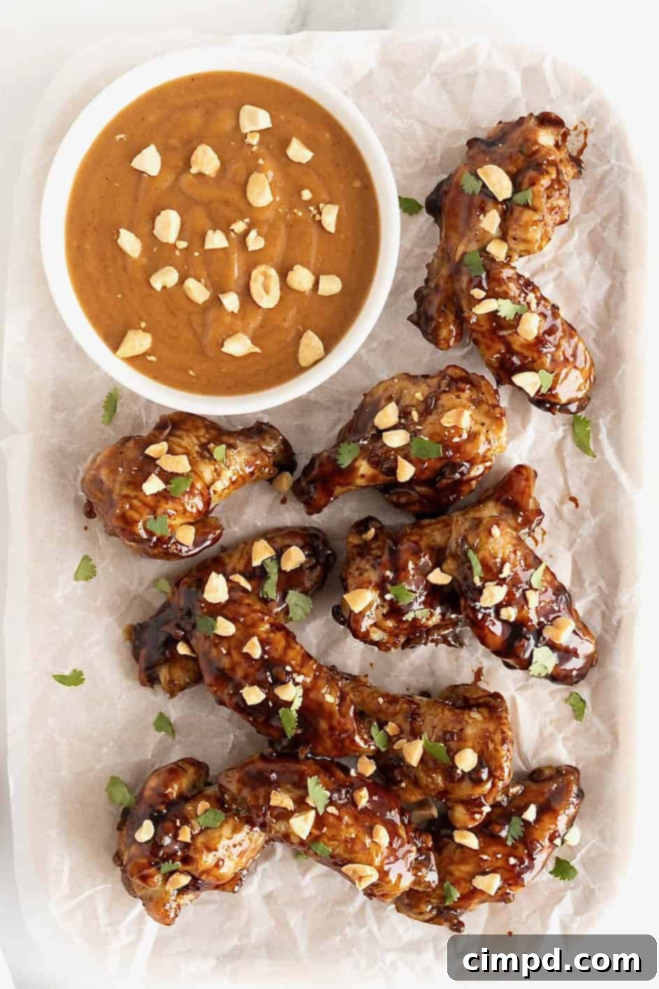 A white serving platter showcasing a pile of golden-brown Baked Crispy Asian Chicken Wings, expertly glazed and garnished, next to a small bowl of peanut dipping sauce.