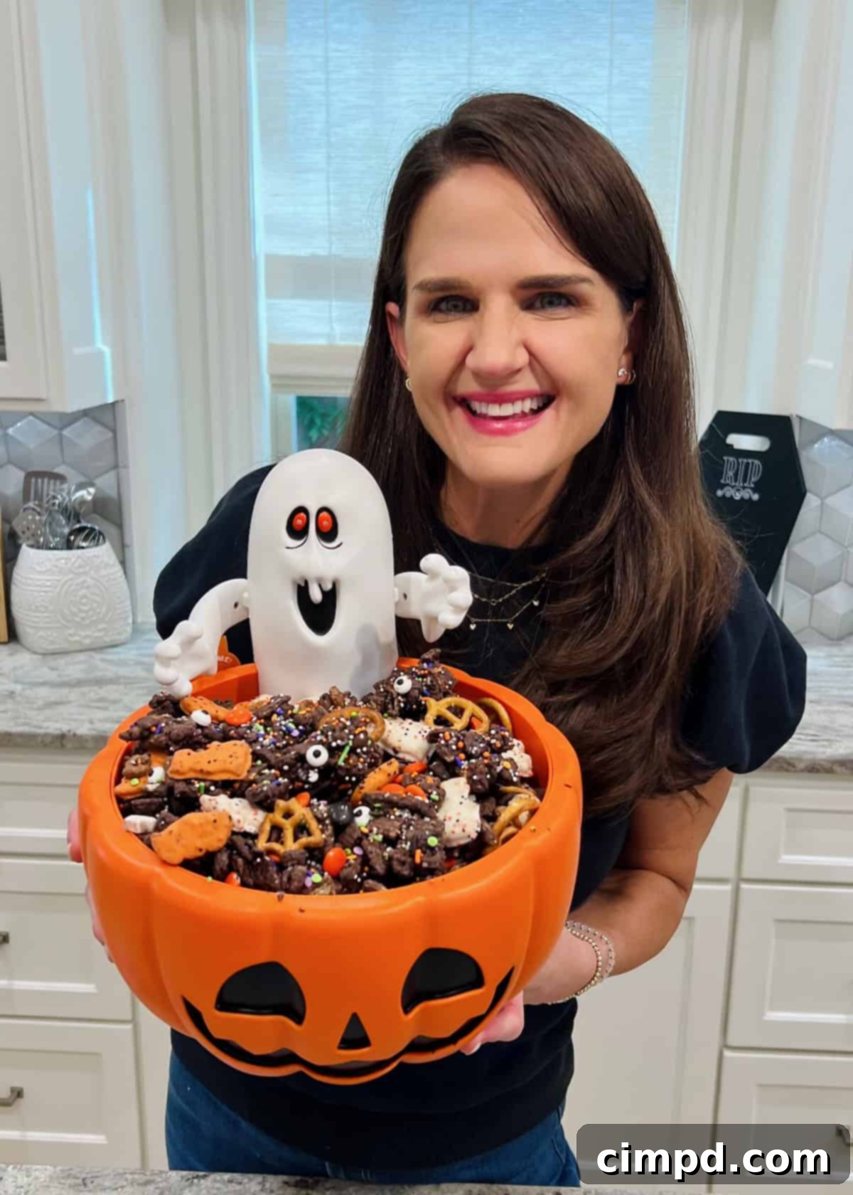 Maegan Brown, the BakerMama, holding a vibrant orange Jack O' Lantern dish overflowing with the delightful Halloween Snack Mix.