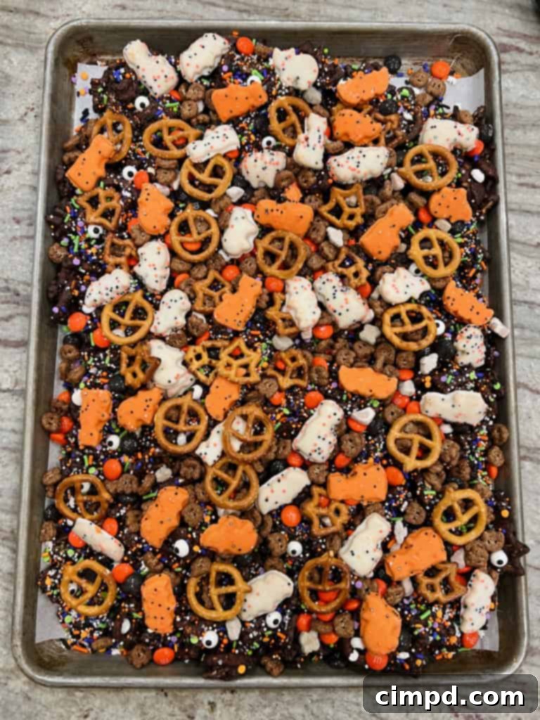 A large pile of freshly prepared Halloween snack mix, brimming with chocolate, pretzels, and candies, on a rimmed baking sheet.