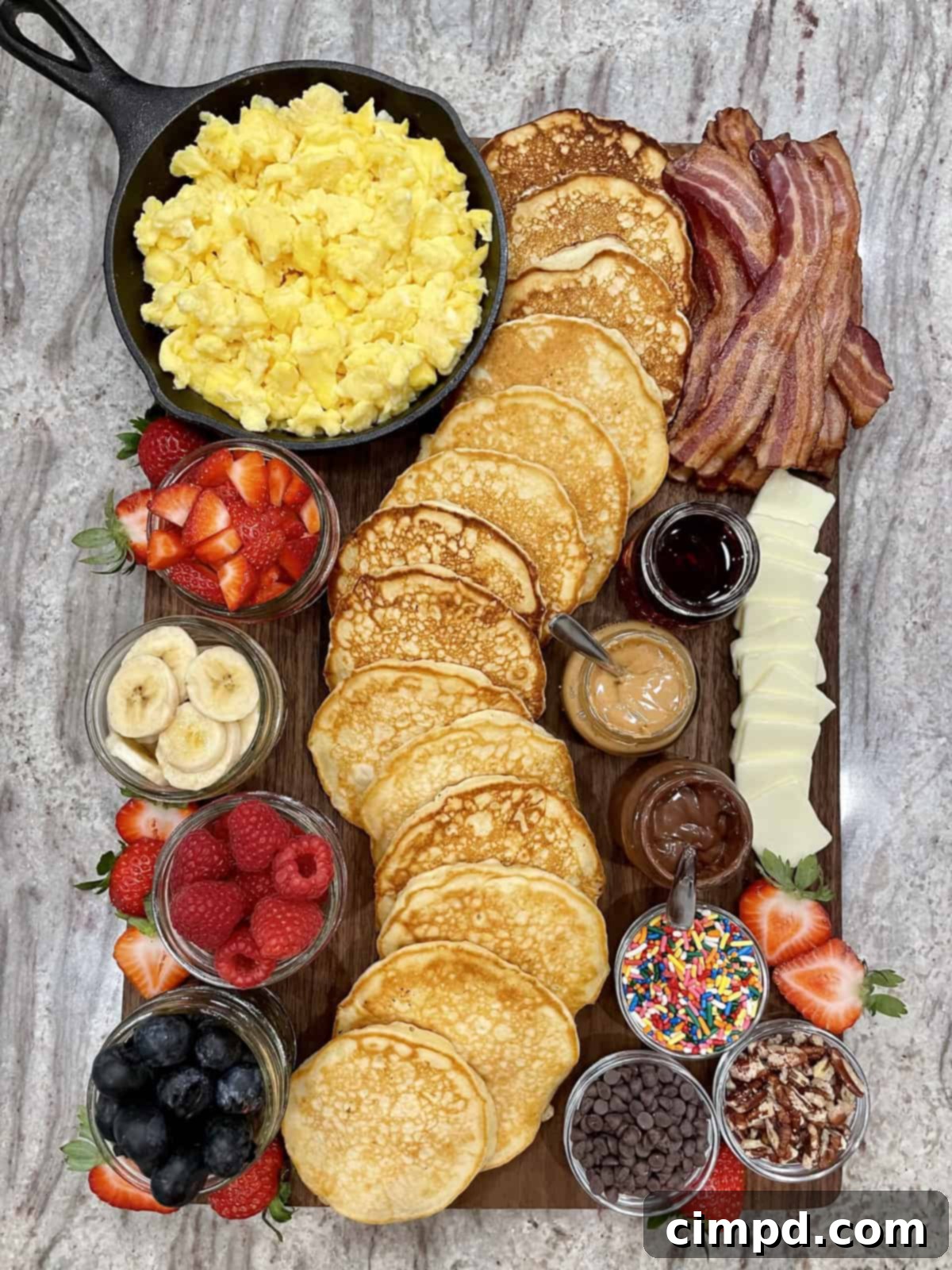 A close-up view of a vibrant Pancake Board, showcasing fluffy pancakes, crispy bacon, an assortment of berries, and various sweet spreads ready for customization.