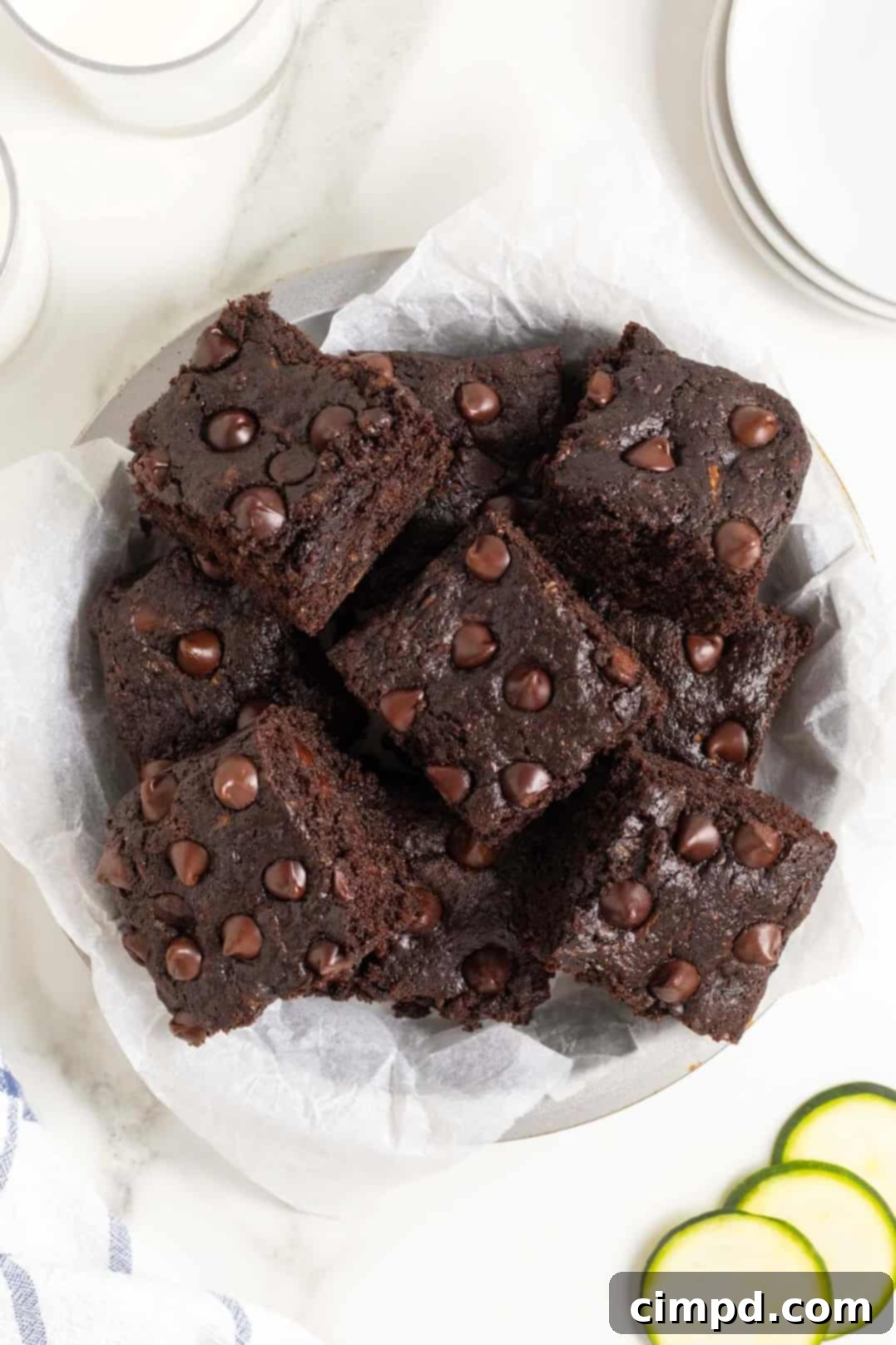 A white plate piled high with rich, dark chocolate zucchini brownies, arranged on a pristine white marble counter. Several thin, fresh slices of vibrant green zucchini are placed beside the brownies, hinting at the secret ingredient.