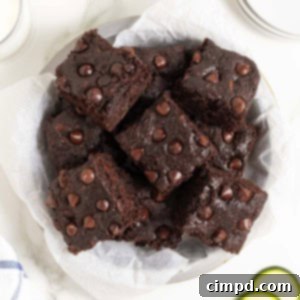 A white plate piled with dark chocolate zucchini brownies on a white marble counter next to several thin slices of zucchini.