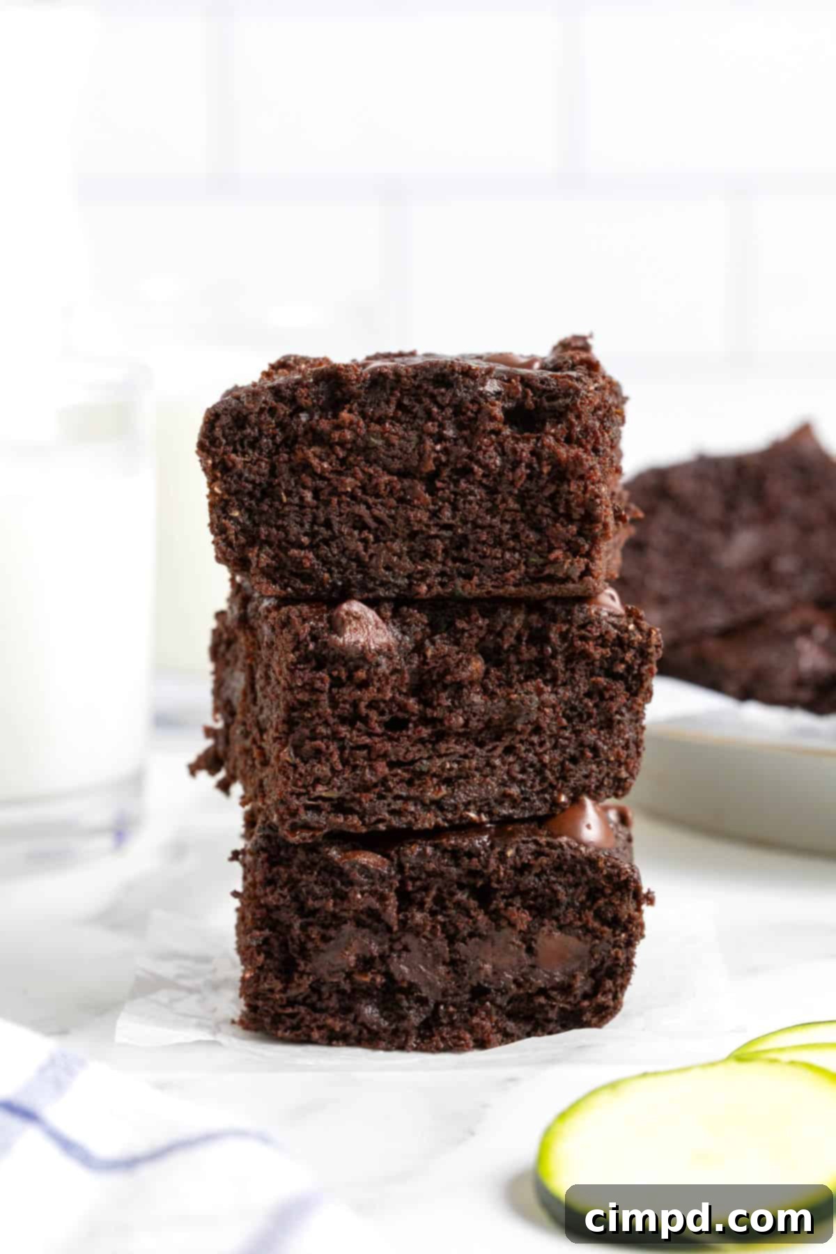 A tempting stack of three dark chocolate zucchini brownies rests on a cool white marble counter. In the soft-focused background, a glass of refreshing milk stands ready, completing the classic brownie pairing.