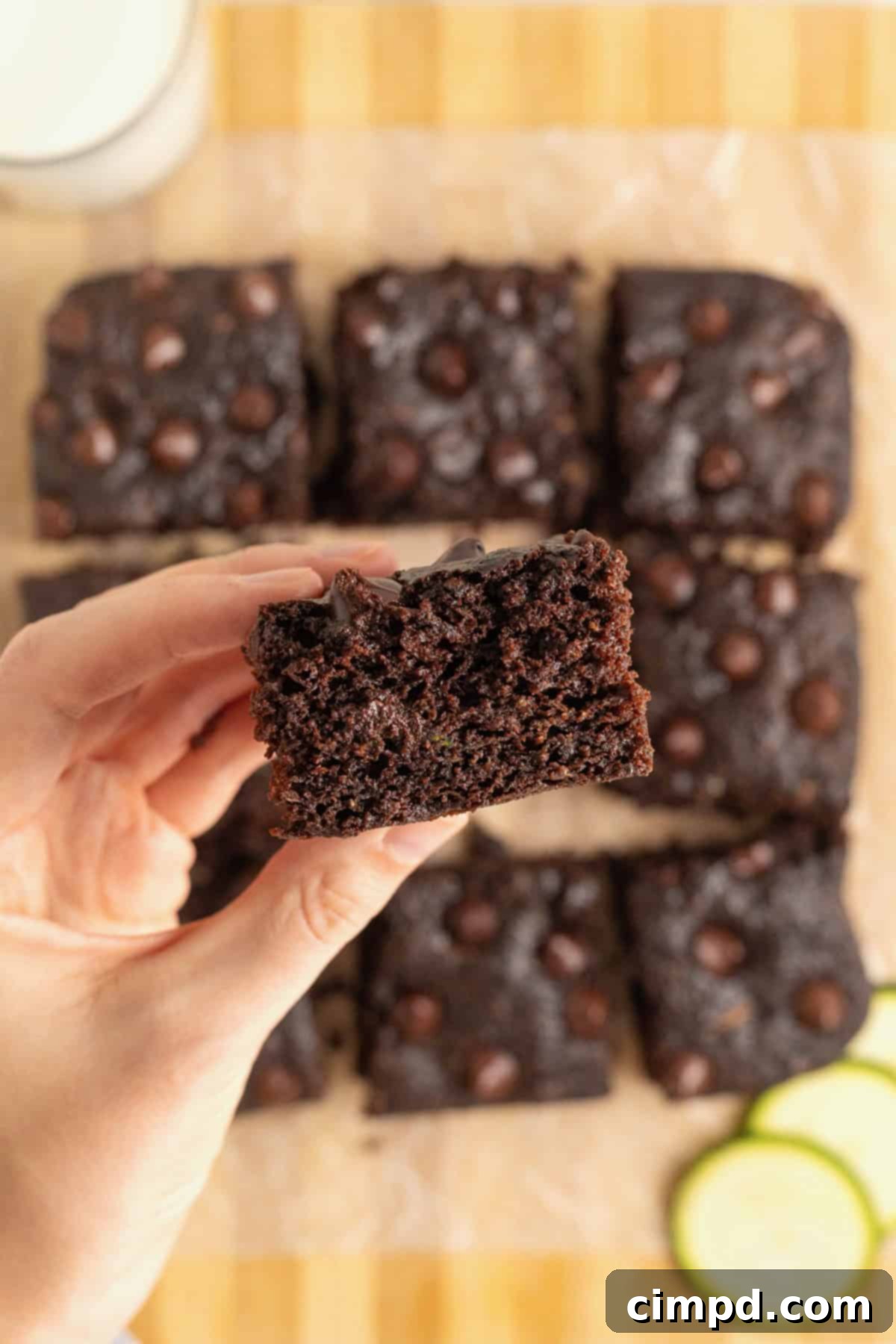 A human hand gracefully reaches for a dark chocolate zucchini brownie from a light wood cutting board, which is laden with several other delicious brownies, inviting a taste.