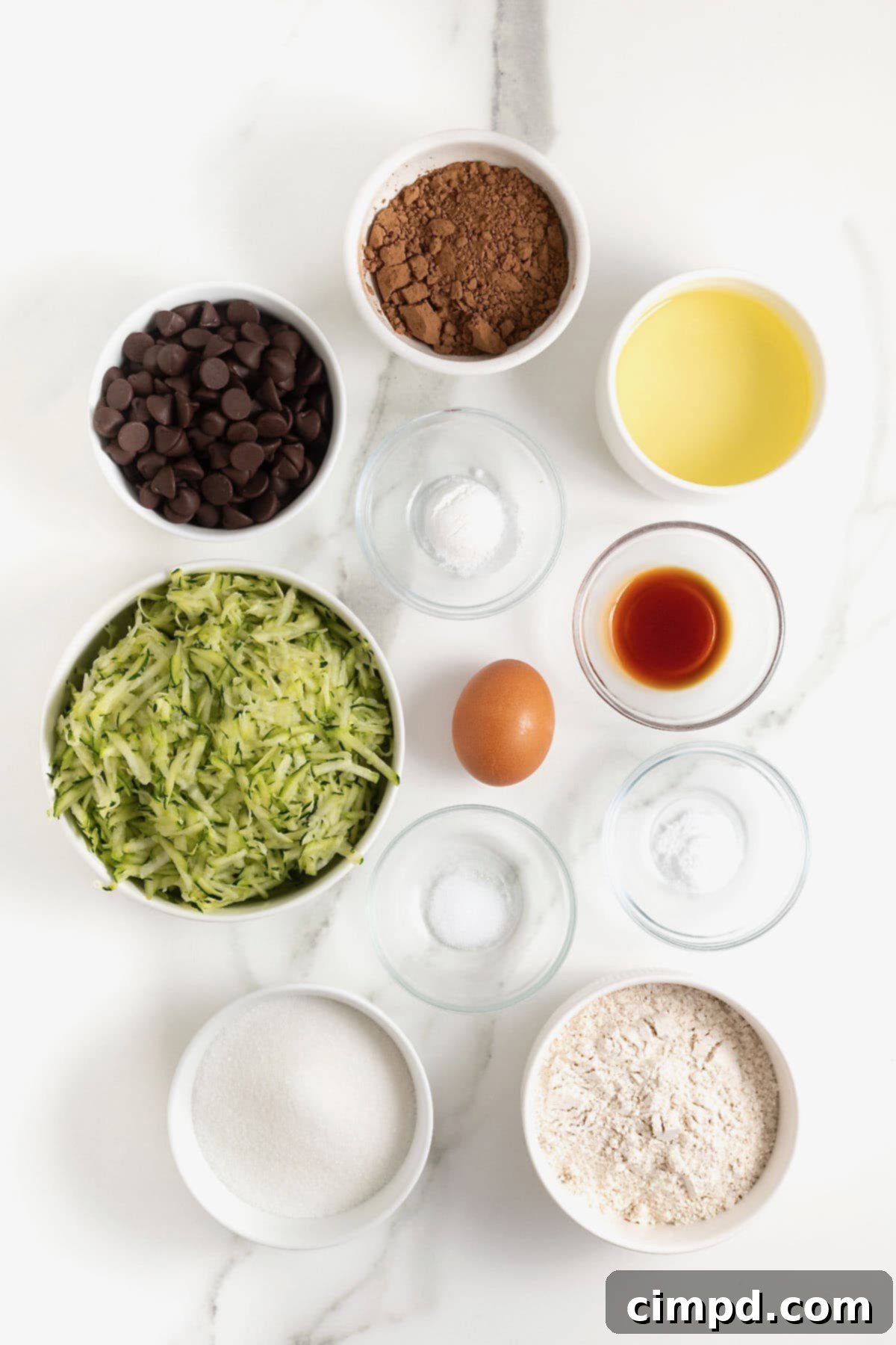 A beautiful flat lay of all the essential ingredients for making zucchini brownies, meticulously arranged in small glass dishes on a pristine white marble counter, ready for baking.