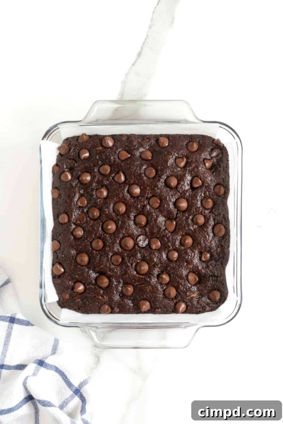 A gleaming square glass baking dish filled with freshly baked brownies, their dark, rich surface studded with glistening melted chocolate chips, just out of the oven.